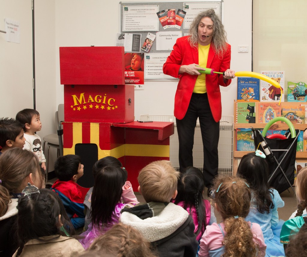 Magician in bright red jacket and yellow t-shirt does magic trick with bike pump and yellow balloon standing in front of children seated in a classroom.